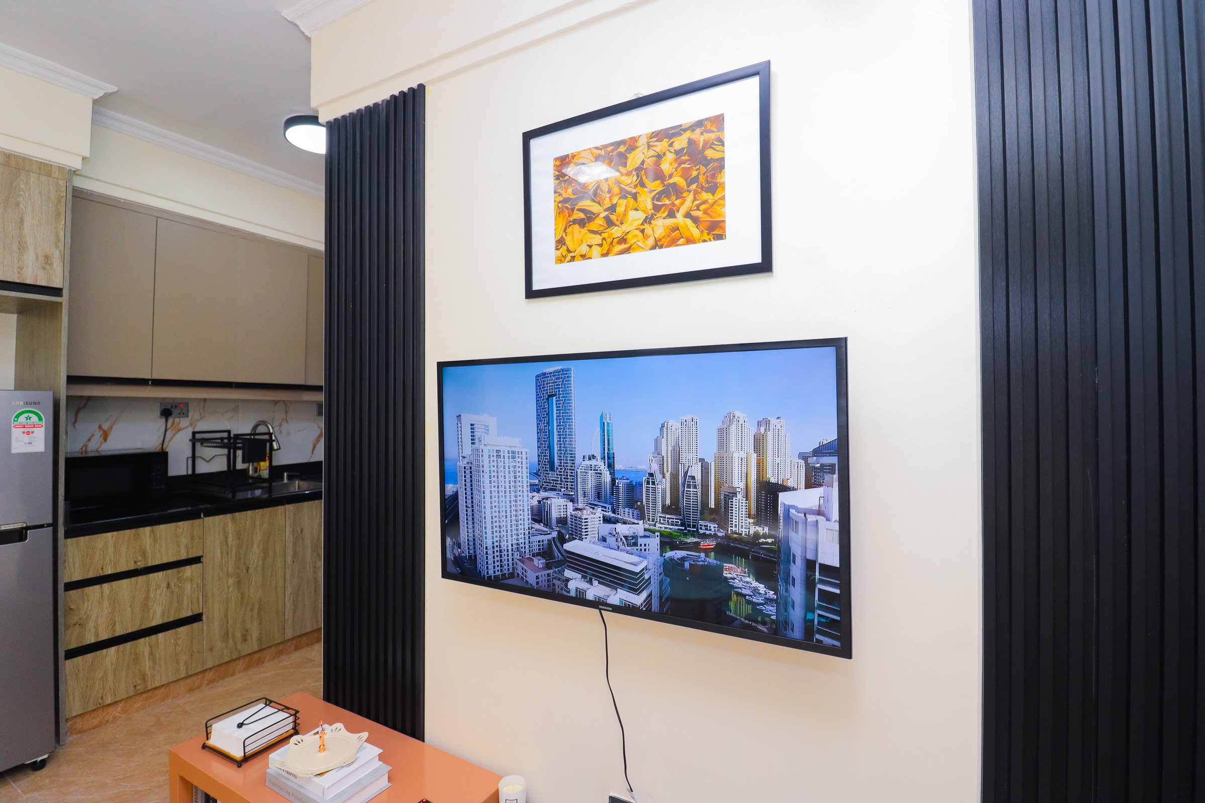 Living room with a wall-mounted TV showing a cityscape, a framed picture above, and a kitchen in the background.
