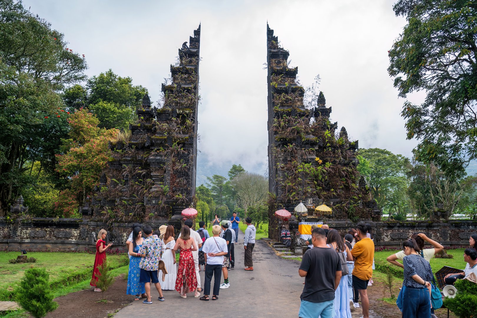 Bali, Indonesia - November 29, 2023: Iconic Balinese temple gate with intricate stone carvings and lush vegetation, surrounded by tourists capturing scenic views against mist-covered mountains.