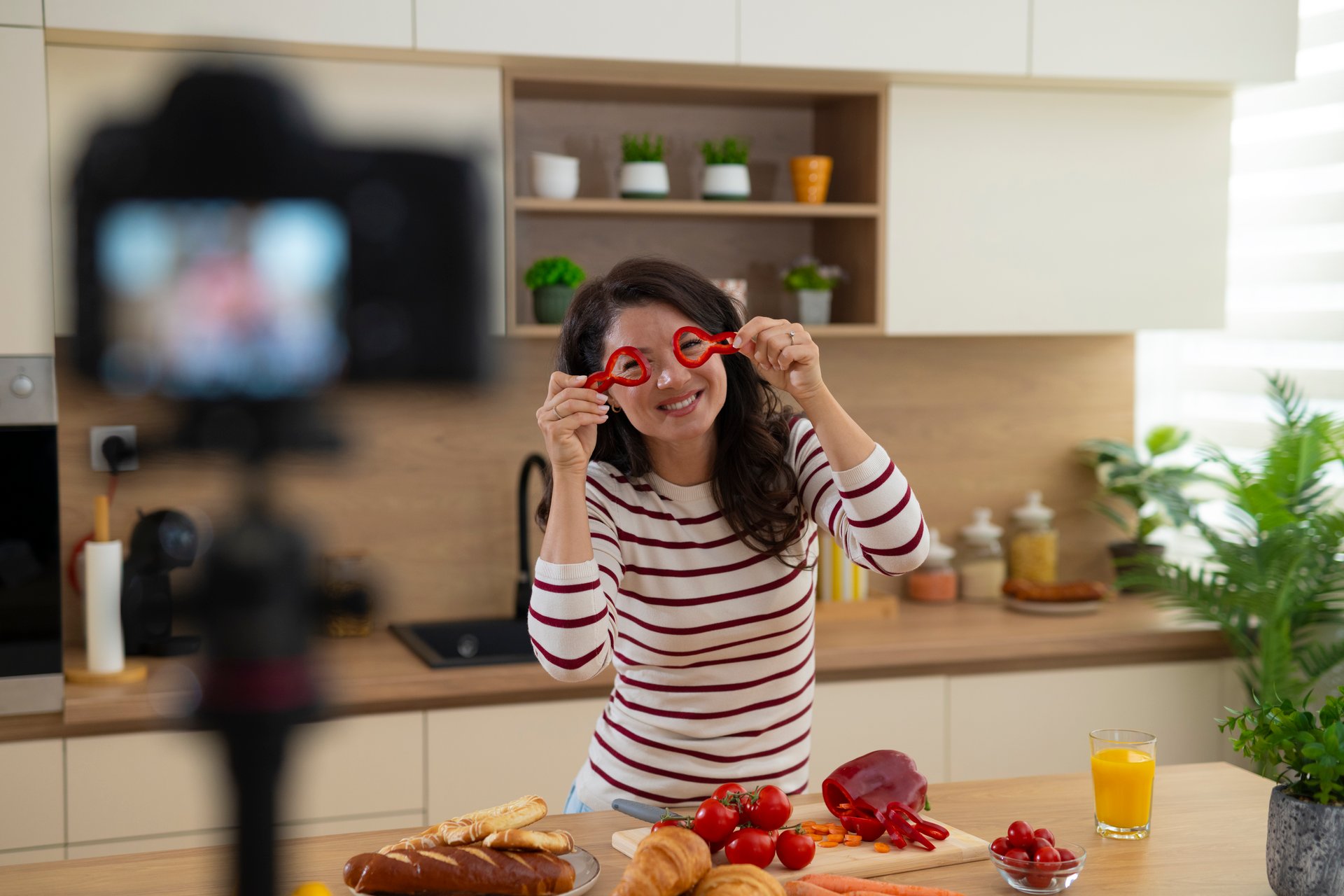 Woman holding red bell peppers playfully while filming a cooking video in kitchen