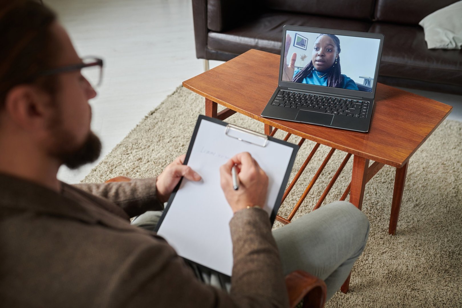 High angle veiw of psychologist making notes on paper while talking to patient online on laptop during psychotherapy session