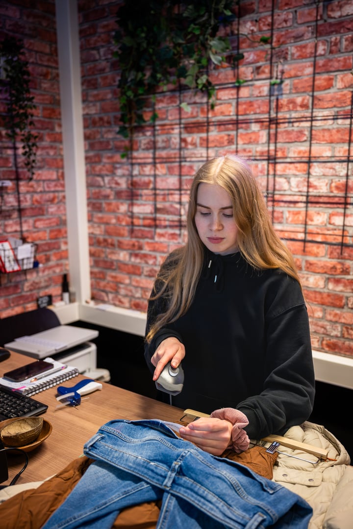 Young woman in black hoodie scanning clothes at a checkout counter in a fashion store, with a brick wall background, concept of retail work