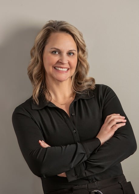 Smiling woman with wavy blonde hair in a black shirt, arms crossed, standing against a neutral wall.