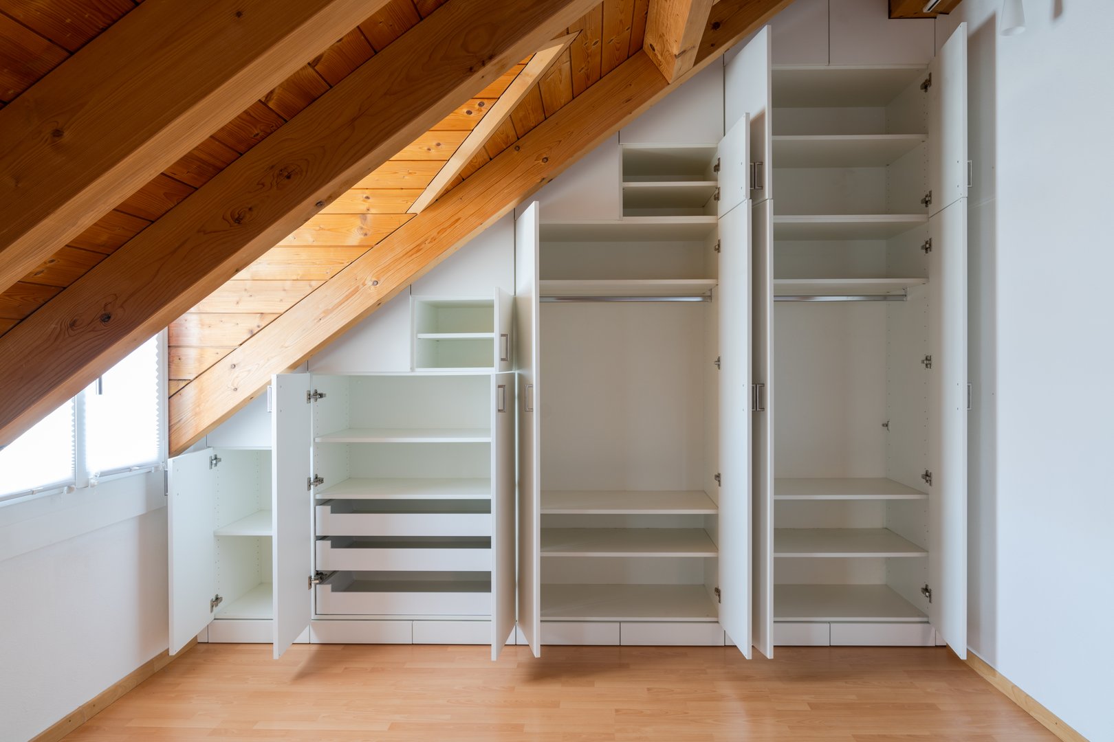 A interior view of a custom-made closet with open doors built into a master bedroom with a sloping roof