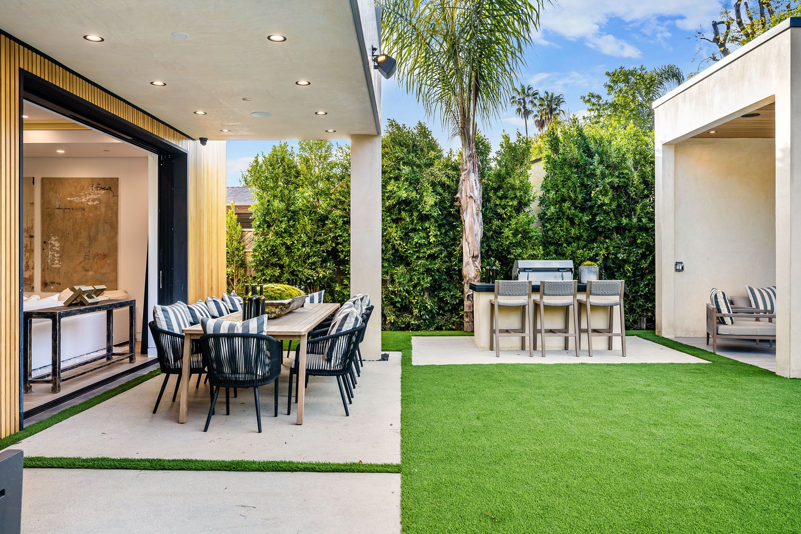 Modern outdoor dining and seating area with a barbecue grill, surrounded by greenery and a clear sky.
