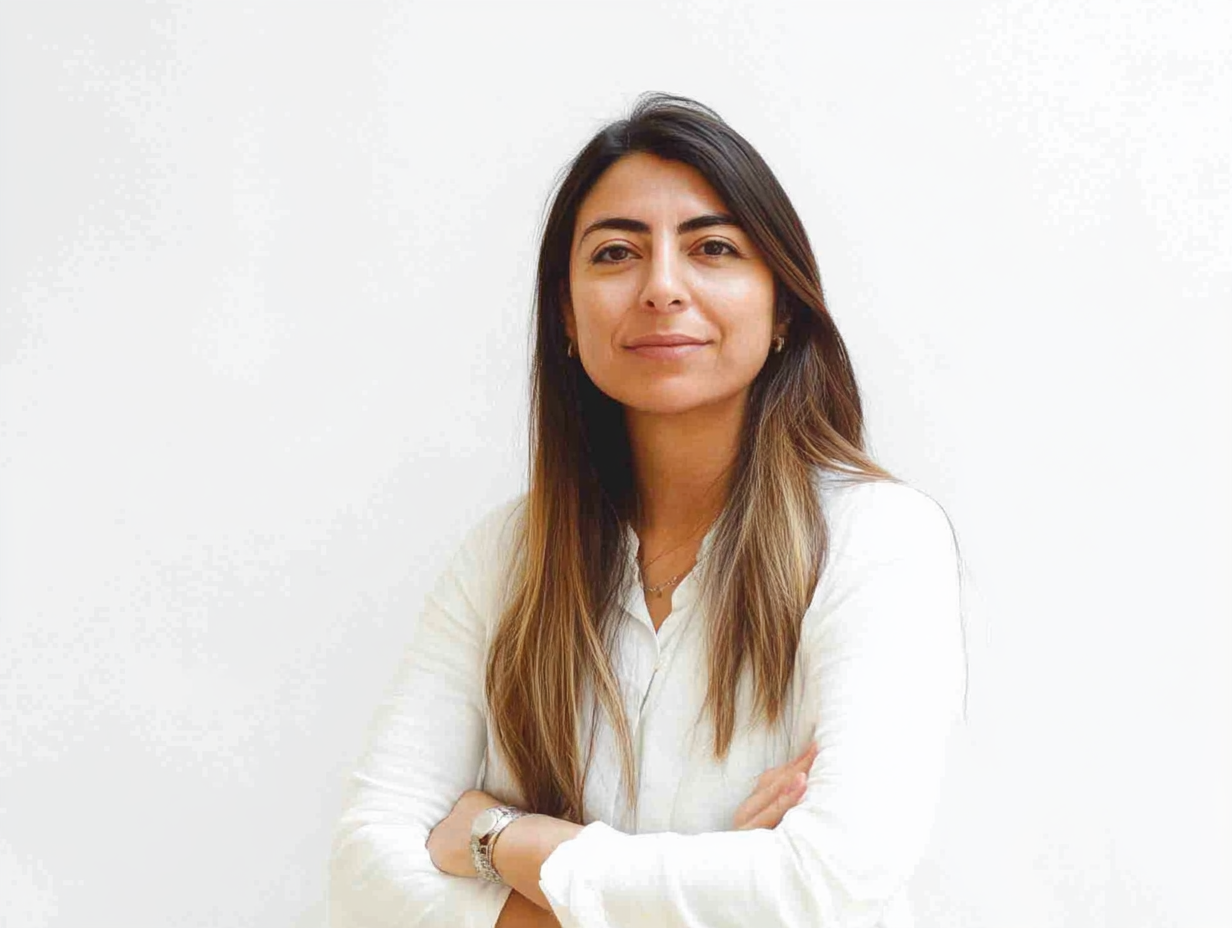 A woman in a white shirt with long brown hair stands against a white background, arms crossed, looking confidently at the camera.