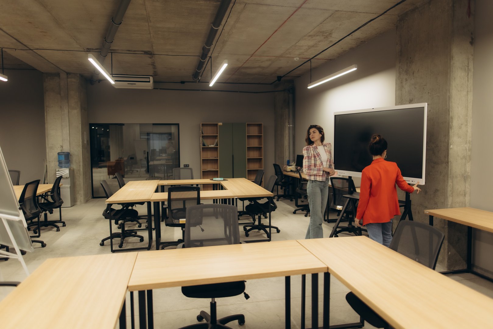 Two women engaging in a discussion in a modern and minimalist open workspace featuring desks, chairs, and a large digital screen, showcasing a collaborative office environment.