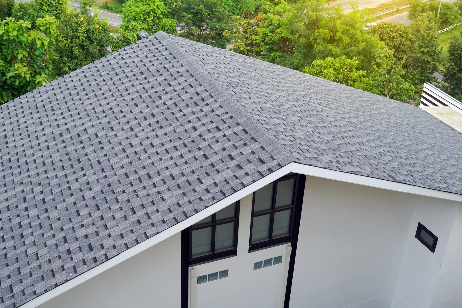 High-angle view of a modern house's steep gray asphalt shingle roof, surrounded by lush green trees