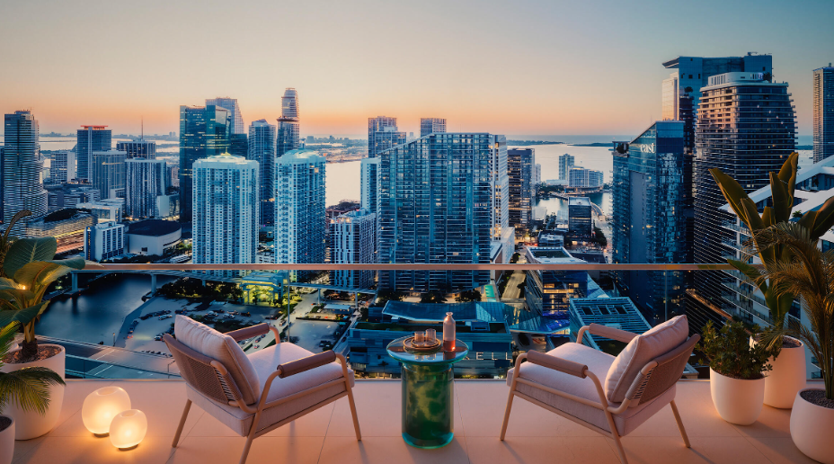 Rooftop terrace view of a city skyline at sunset, with two chairs, a table, and potted plants overlooking skyscrapers and water.