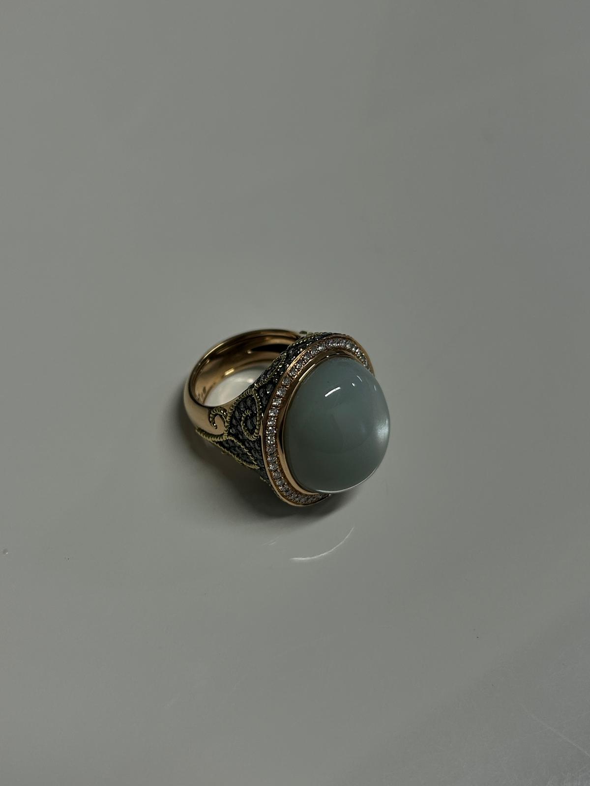 A gold ring with an ornate band and a large, smooth gray gemstone set in the center, surrounded by small diamonds.