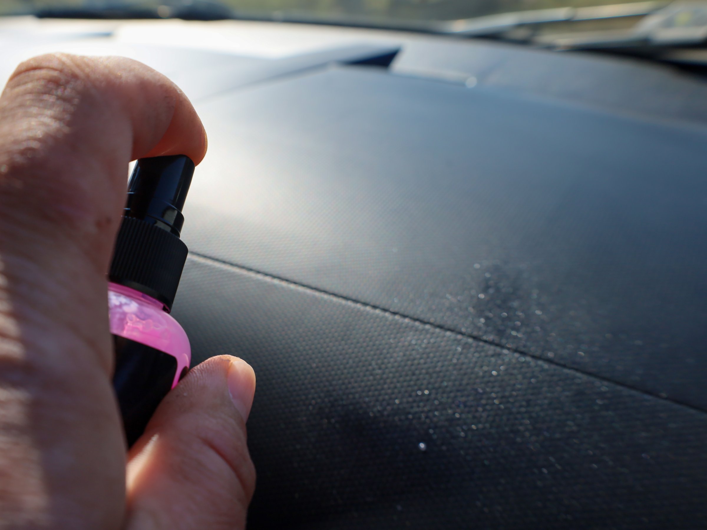 Close-up of a hand spraying cleaner on a car dashboard. Small particles of dust and dirt are visible settling on the surface. A person is using a spray to clean and refresh the car's interior.