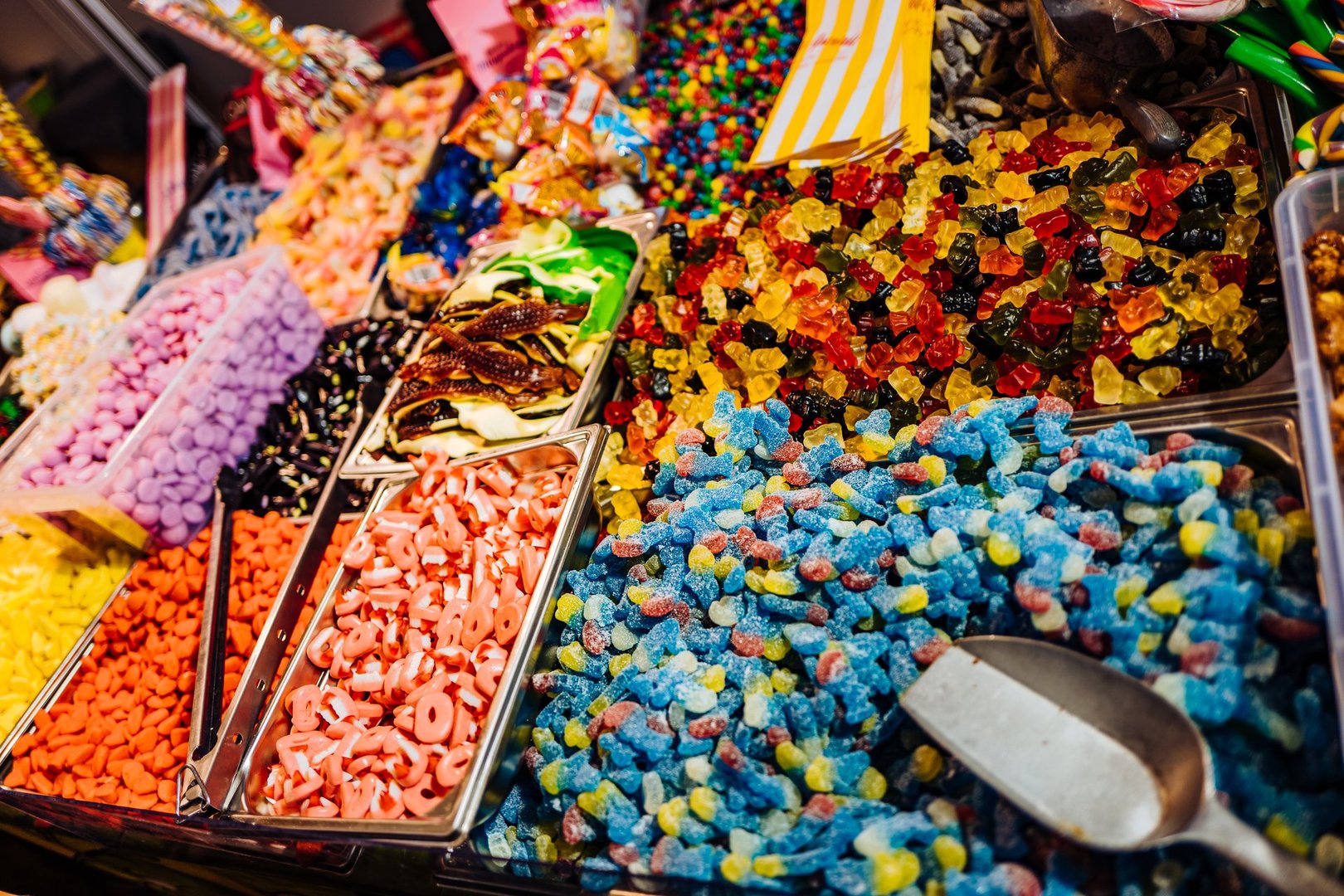 Assorted colorful candies displayed in trays at a market, including gummy bears, sour candies, and chocolate.