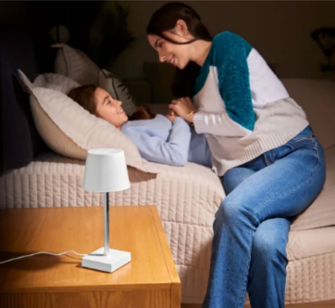 A woman leans over a young girl lying on a sofa, both smiling, with a small white lamp on a nearby table.