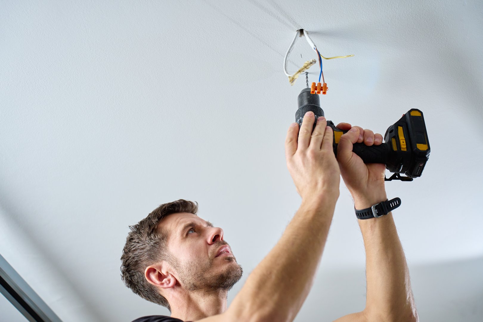 Man wearing work overalls drills hole in ceiling using cordless power drill in indoor room. Concept of home renovation, installation and professional maintenance service.