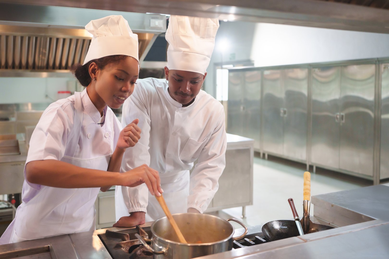 African American Female and Black male chefs in white uniforms and hats stirring pot together, teamwork in modern professional kitchen, culinary training