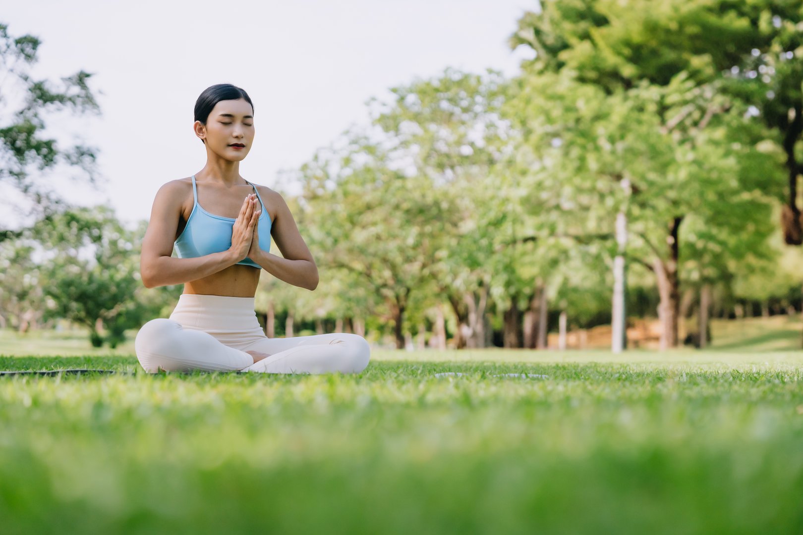 Asian woman meditating in a peaceful park setting, sitting cross-legged on green grass with trees in the background on a sunny day. Concept of mindfulness, self-care, and outdoor relaxation
