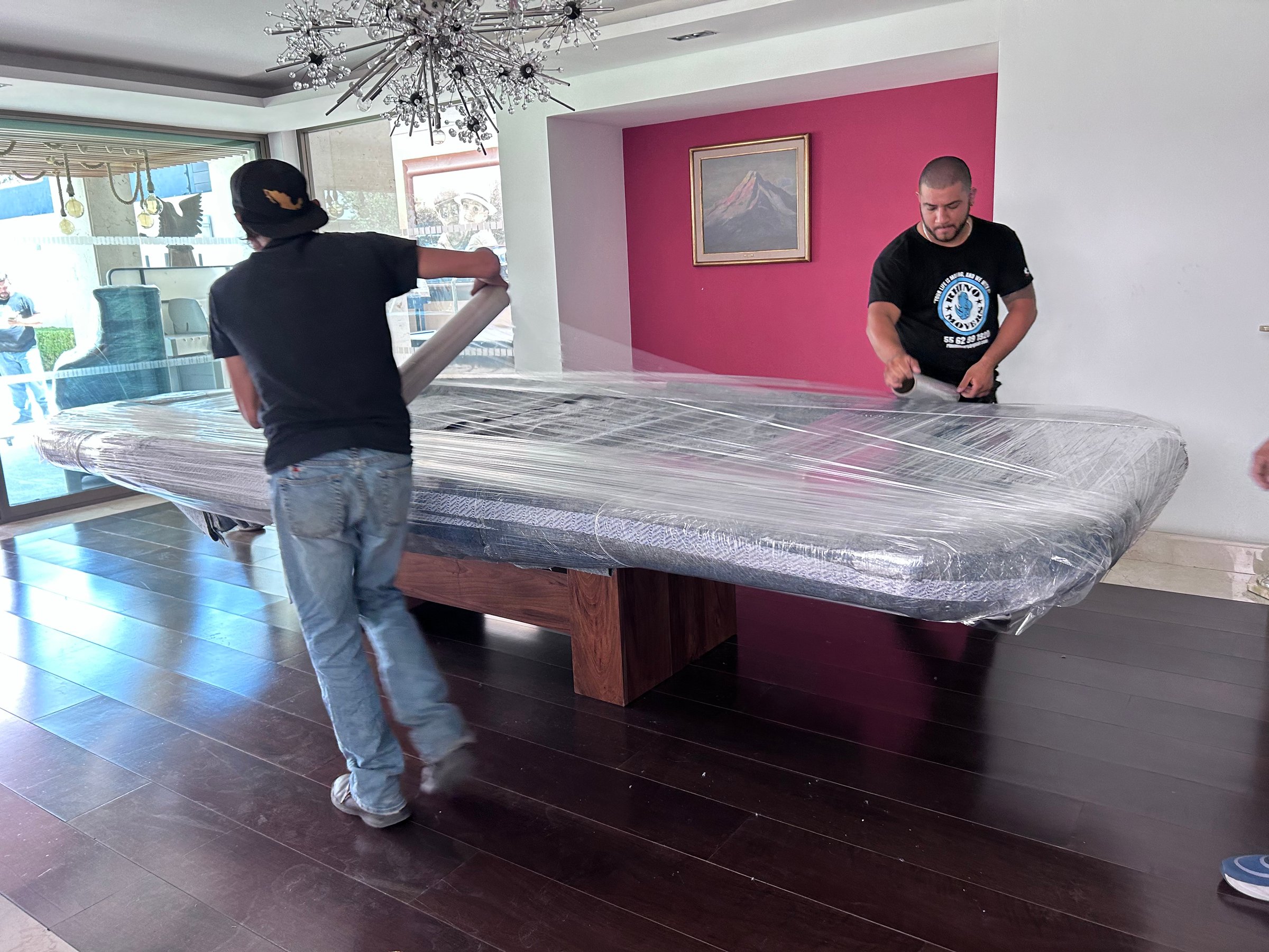 Two men wrapping a large table with plastic in a modern living room with a pink accent wall and a chandelier overhead.