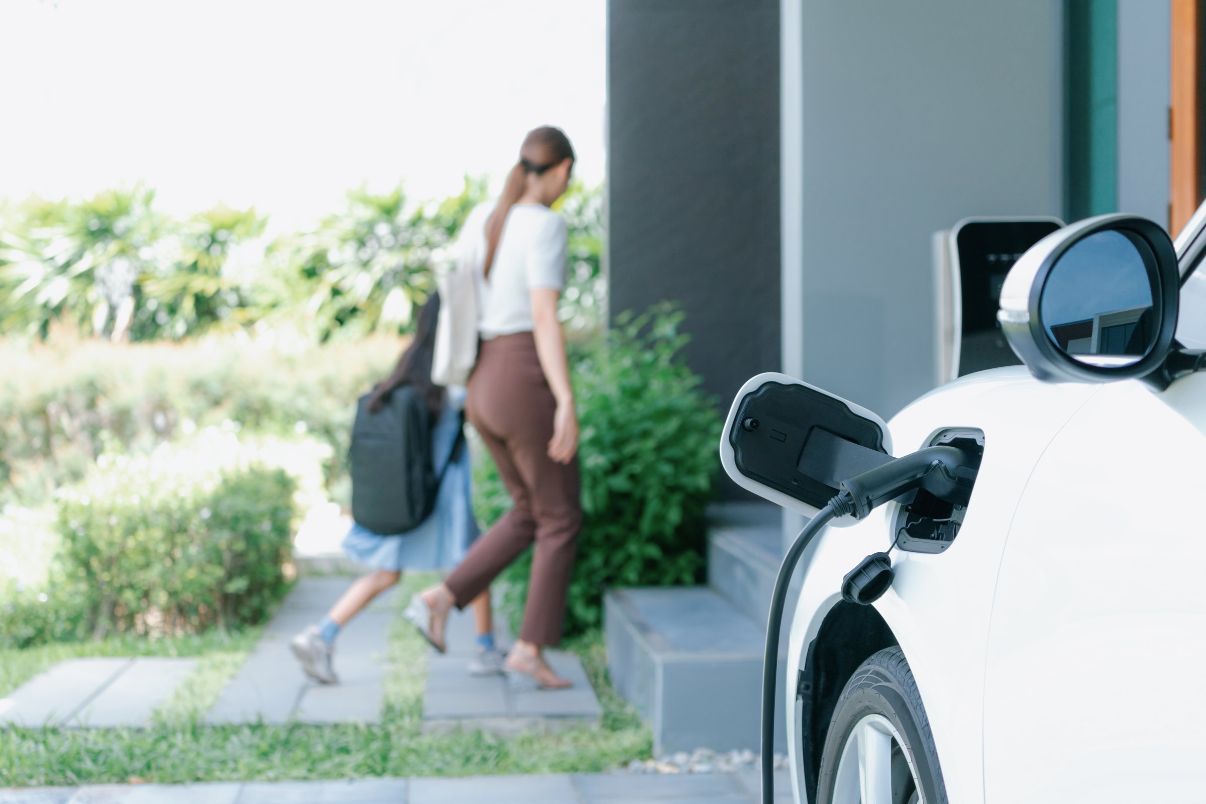 Focus electric vehicle recharging at home charging station plugged in with EV charger device with blurred background of progressive mother and daughter walking as concept for sustainability of energy.