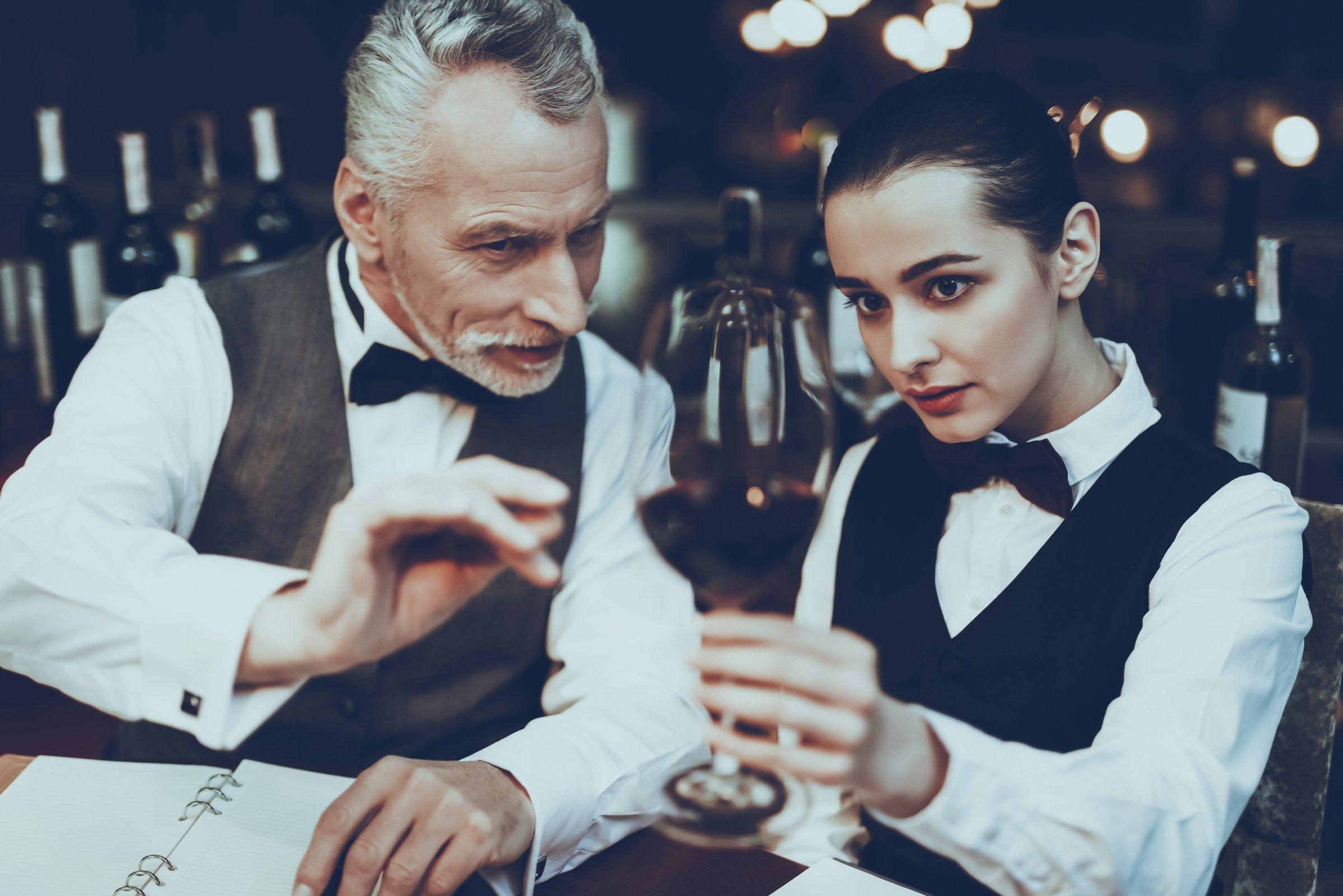 Experienced Sommeliers is Working in Restaurant. Sommeliers is Tasting Red Wine. Sommeliers is Old Man and Young Woman. People is Sitting at Table. People is Wearing Special Uniform.