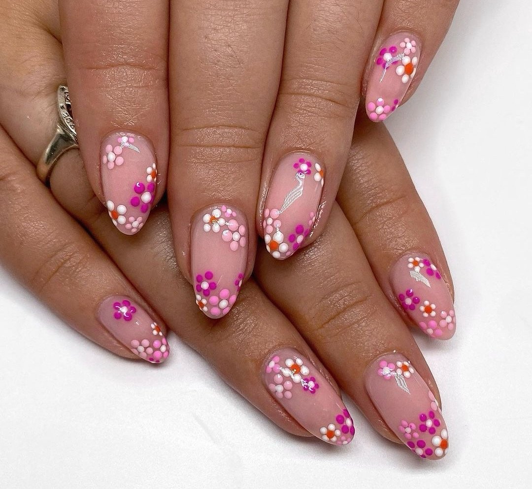 Pink nails with colorful floral designs in orange, pink, and white, featuring small dots and a silver ring on one finger.