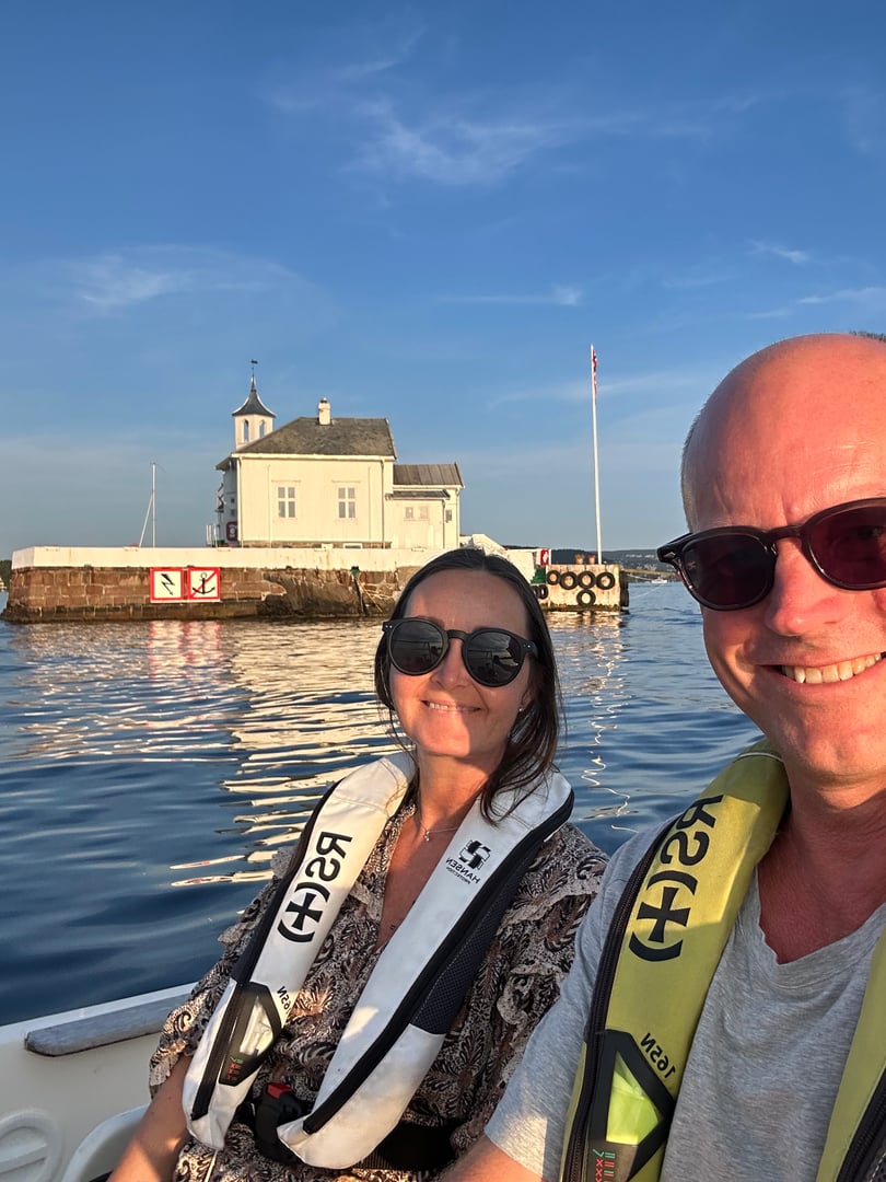 Two people in a boat wearing life jackets, smiling with a small building and calm water in the background.