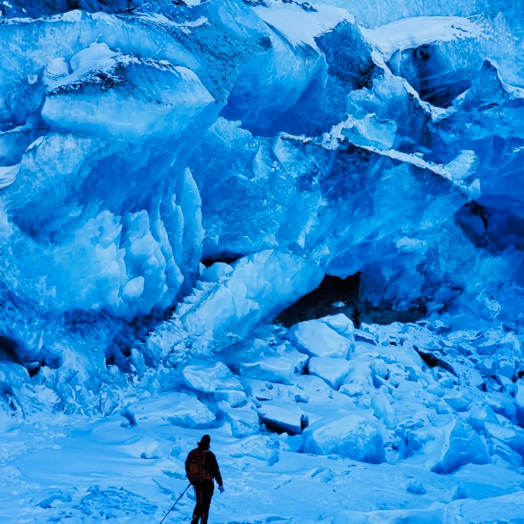 A person with hiking gear stands before a large, icy glacier with deep blue hues and jagged formations.