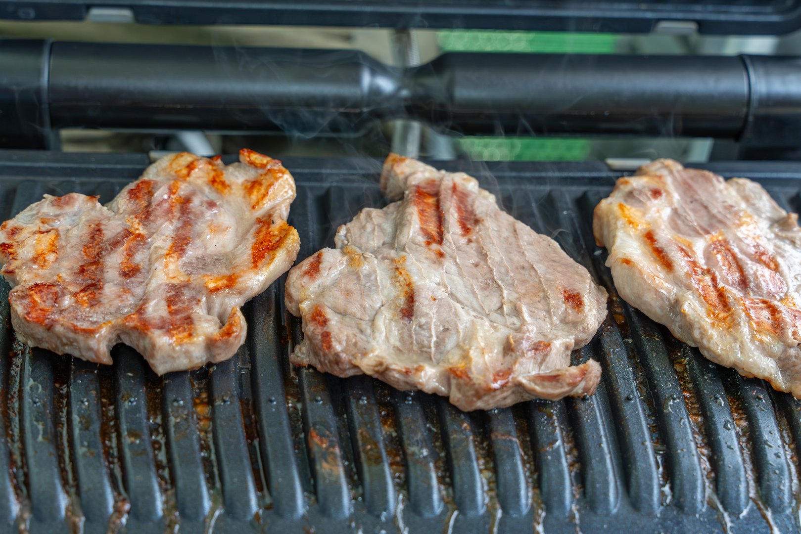 Three juicy pieces of meat are grilling on a contact grill, showing distinct grill marks. Light smoke rises as the meat sizzles.