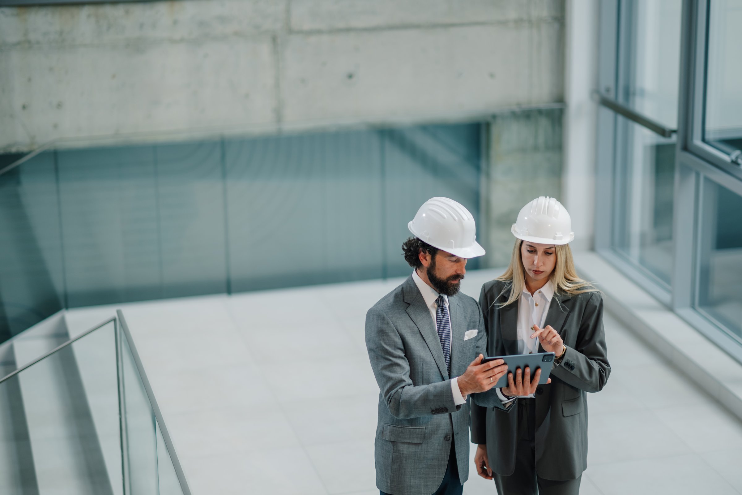 Two architects wearing hardhats are using a digital tablet to discuss blueprints in a modern building