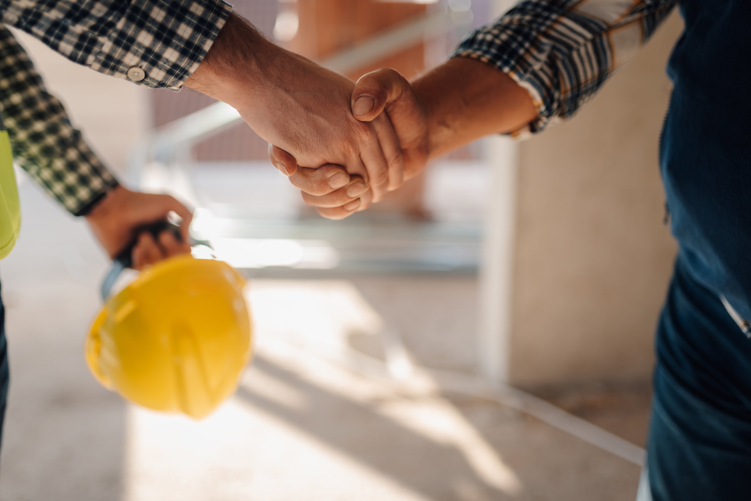 Two construction workers shaking hands on a building site, holding a yellow hardhat, celebrating successful agreement