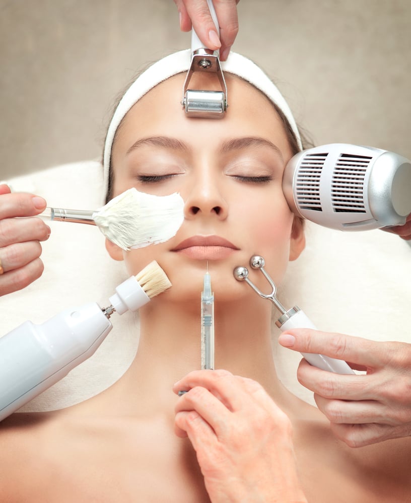 Woman receiving various beauty treatments including facial masks, roller, and syringe on her face.