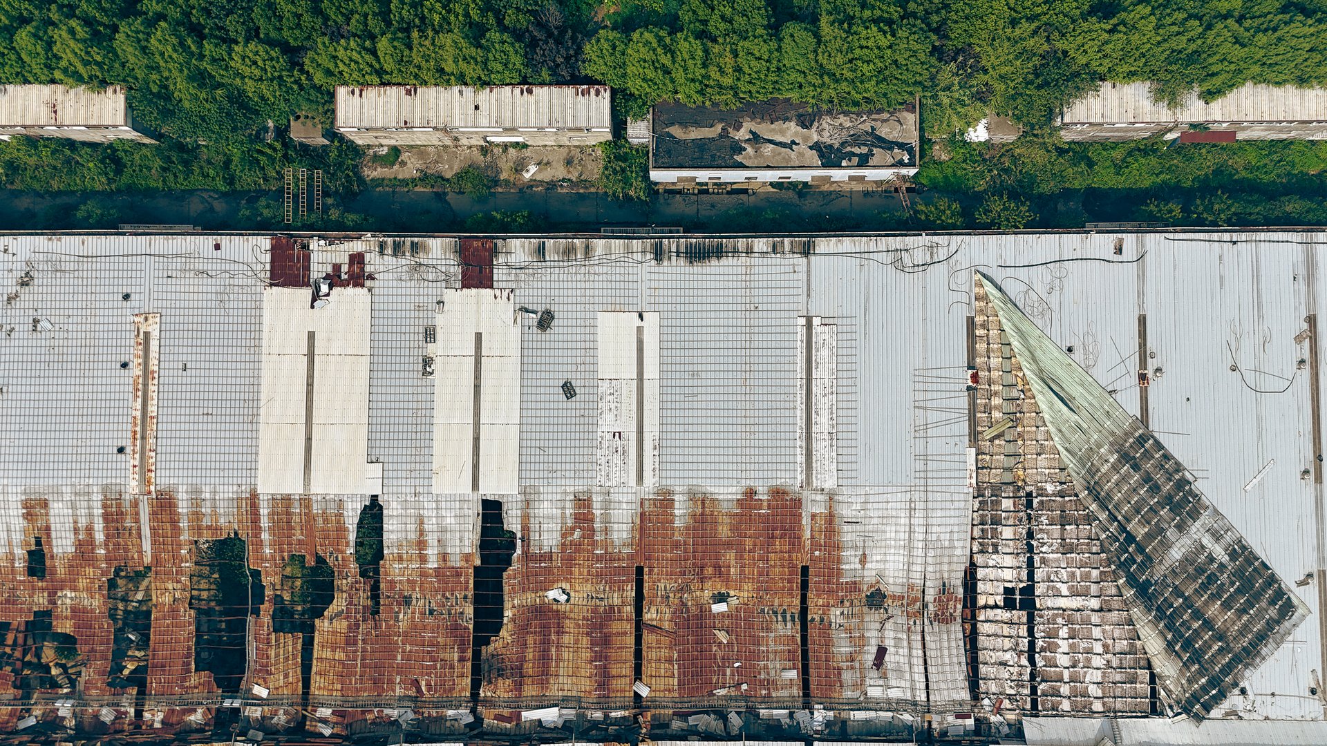 Drone aerial photographs of an abandoned industrial warehouse with rusted corrugated metal roofs, collapsed sections, and visible decay. The site shows overgrown vegetation and signs of long-term neglect, highlighting urban decay and industrial decline.