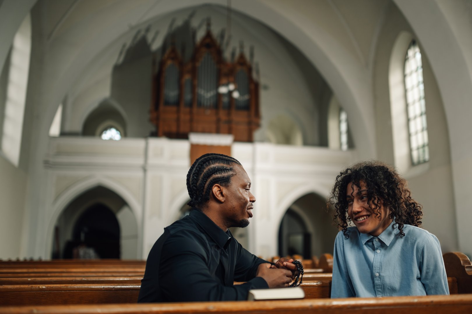 Priest listens attentively to a young man in a church pew, holding rosary beads, exuding faith, spirituality, and guidance in a serene atmosphere
