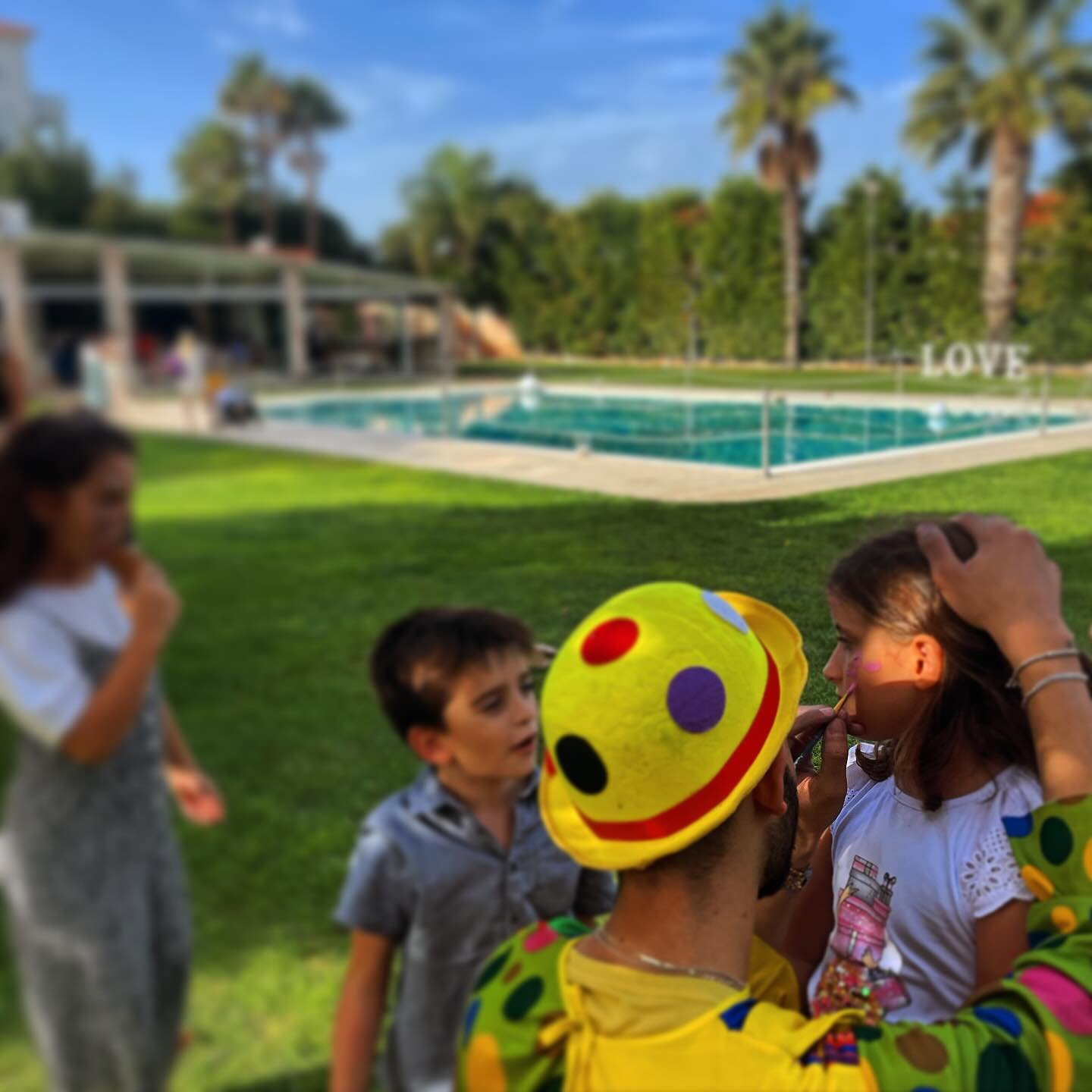 Clown applying face paint to a child near a pool at an outdoor event with palm trees in the background.