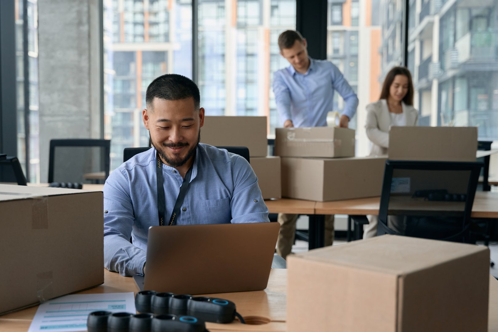 Smiling company manager working on his laptop while his colleagues packing cardboard boxes