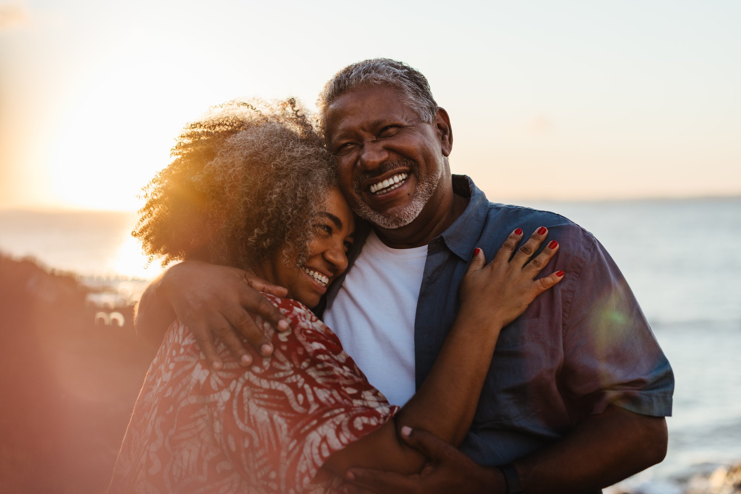Elderly pair embraces at sunset, radiating joy and love by the seaside. A beautiful moment of togetherness and happiness.