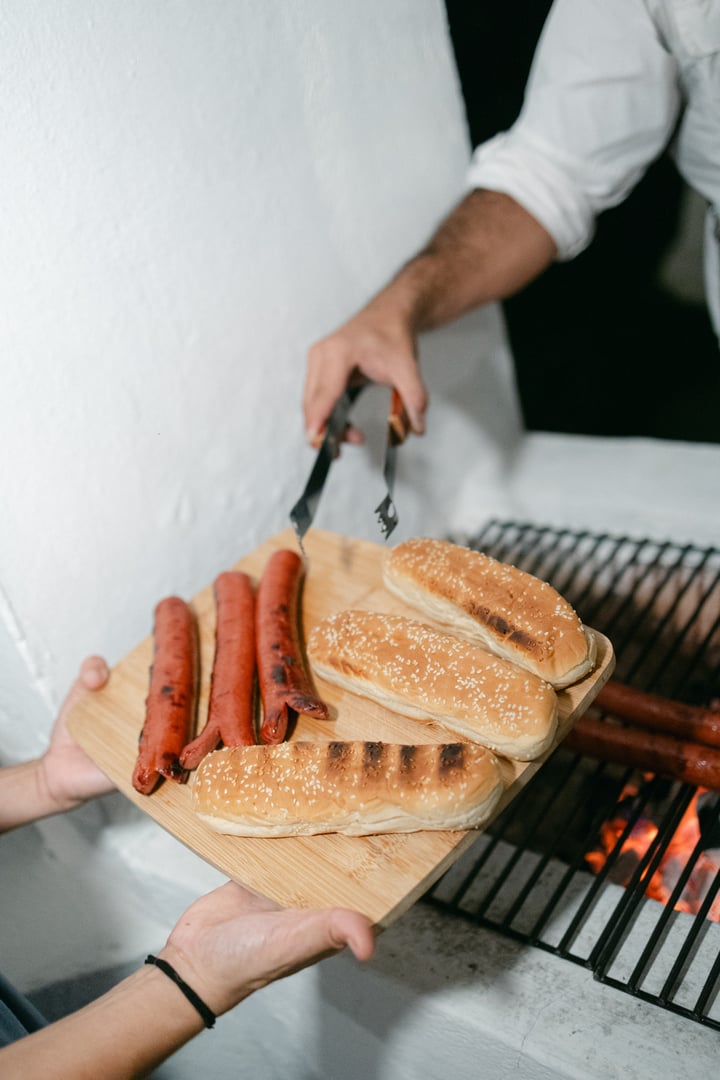 Person using tongs to place grilled hot dogs on buns over a barbecue grill.