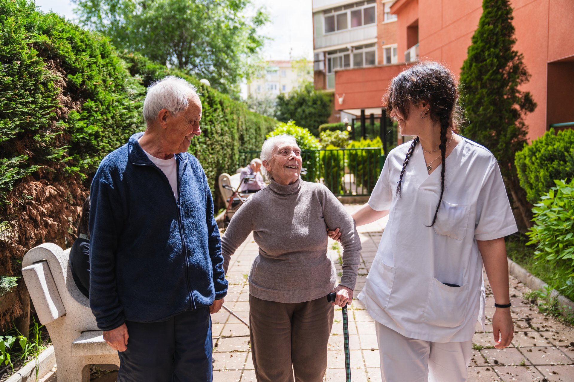 Nurse helping senior woman with walking stick in a sunny nursing home garden, accompanied by a senior man