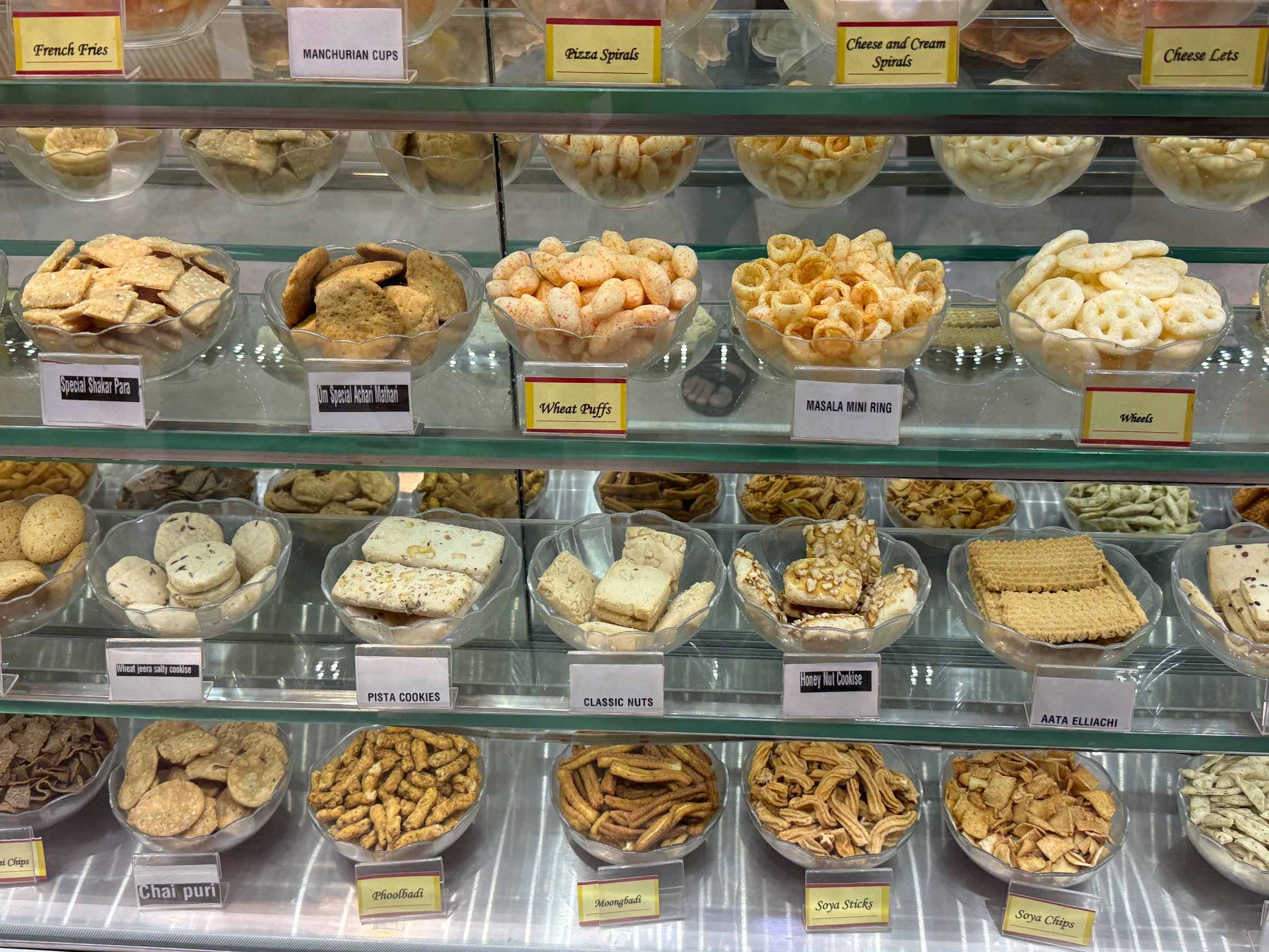 A display of various snacks and treats in a glass case. The snacks are arranged in different bowls and trays, with some labeled as Aloo Gobi and Aloo Paratha. The display is colorful and inviting