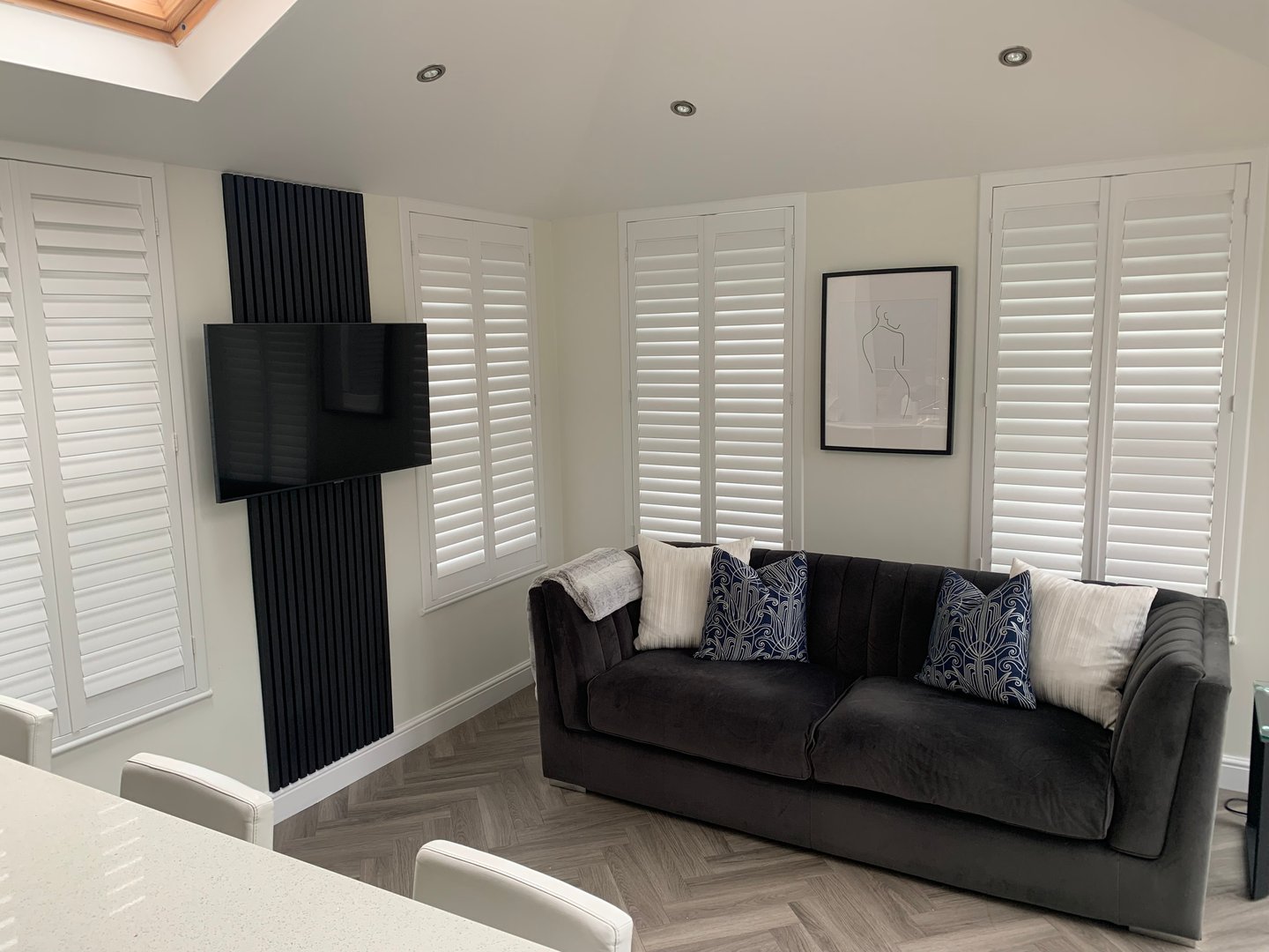 Modern living room with a dark sofa, patterned pillows, wall-mounted TV, white shutters, and minimalist decor.