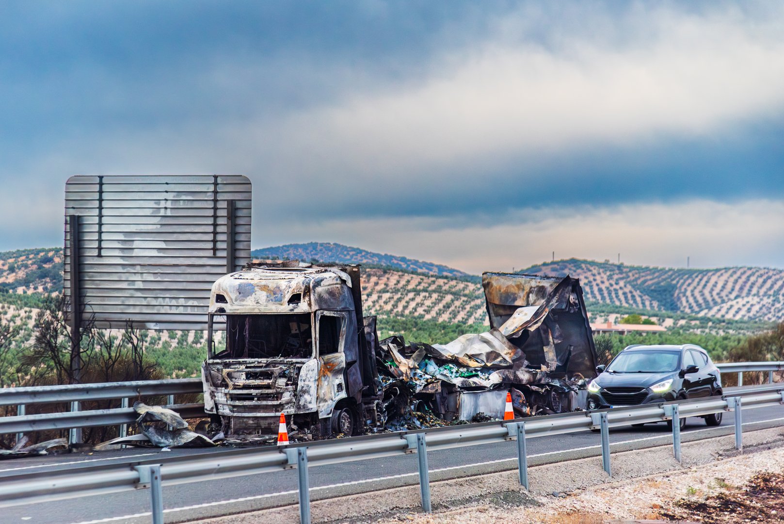 Completely burned-out truck after catching fire on the road, highway with olive fields in the background.