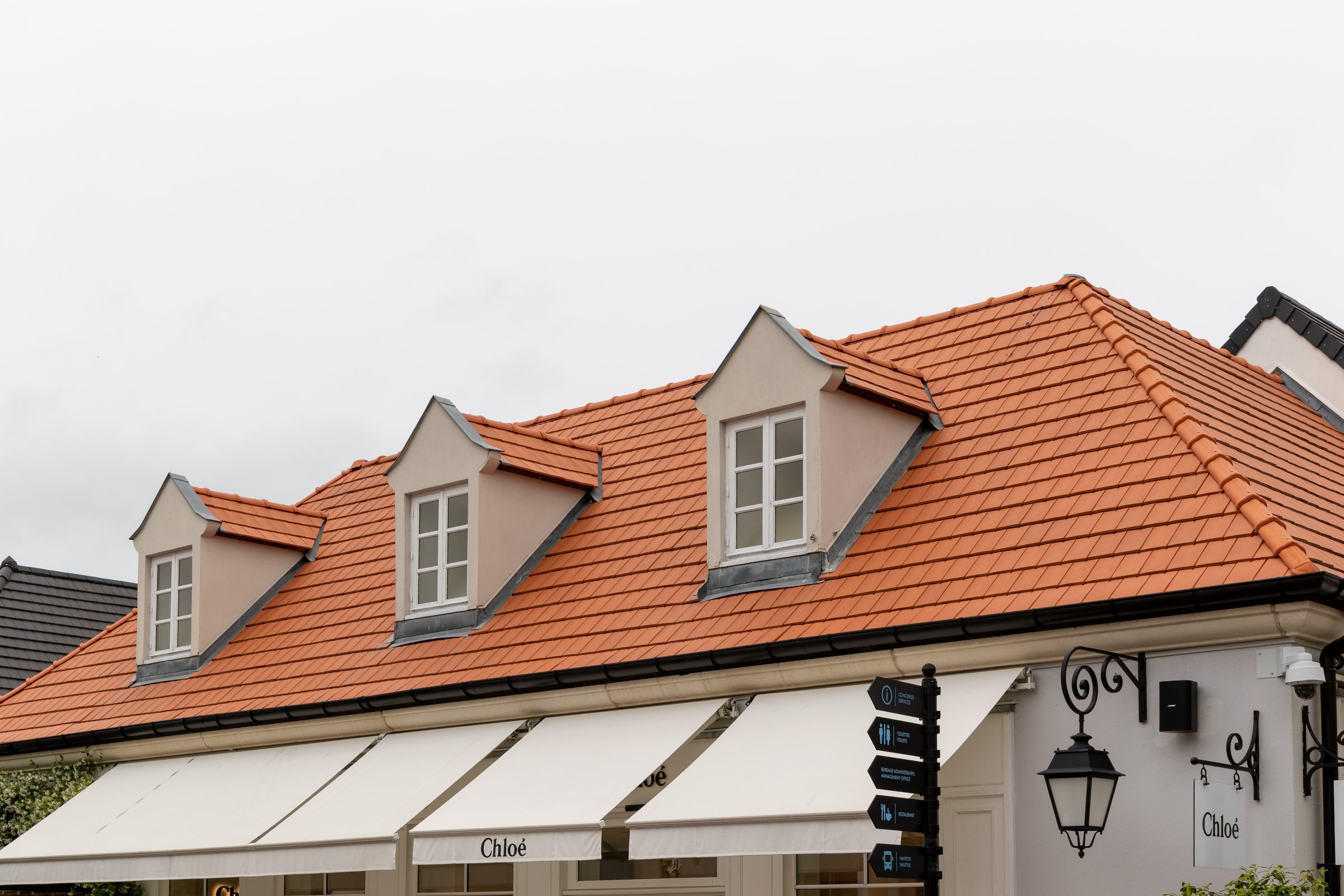 Exterior view of a building with orange tile roof, dormer windows, and white awnings. A Chloe sign and decorative lamp are visible at La Vallee Village, France.