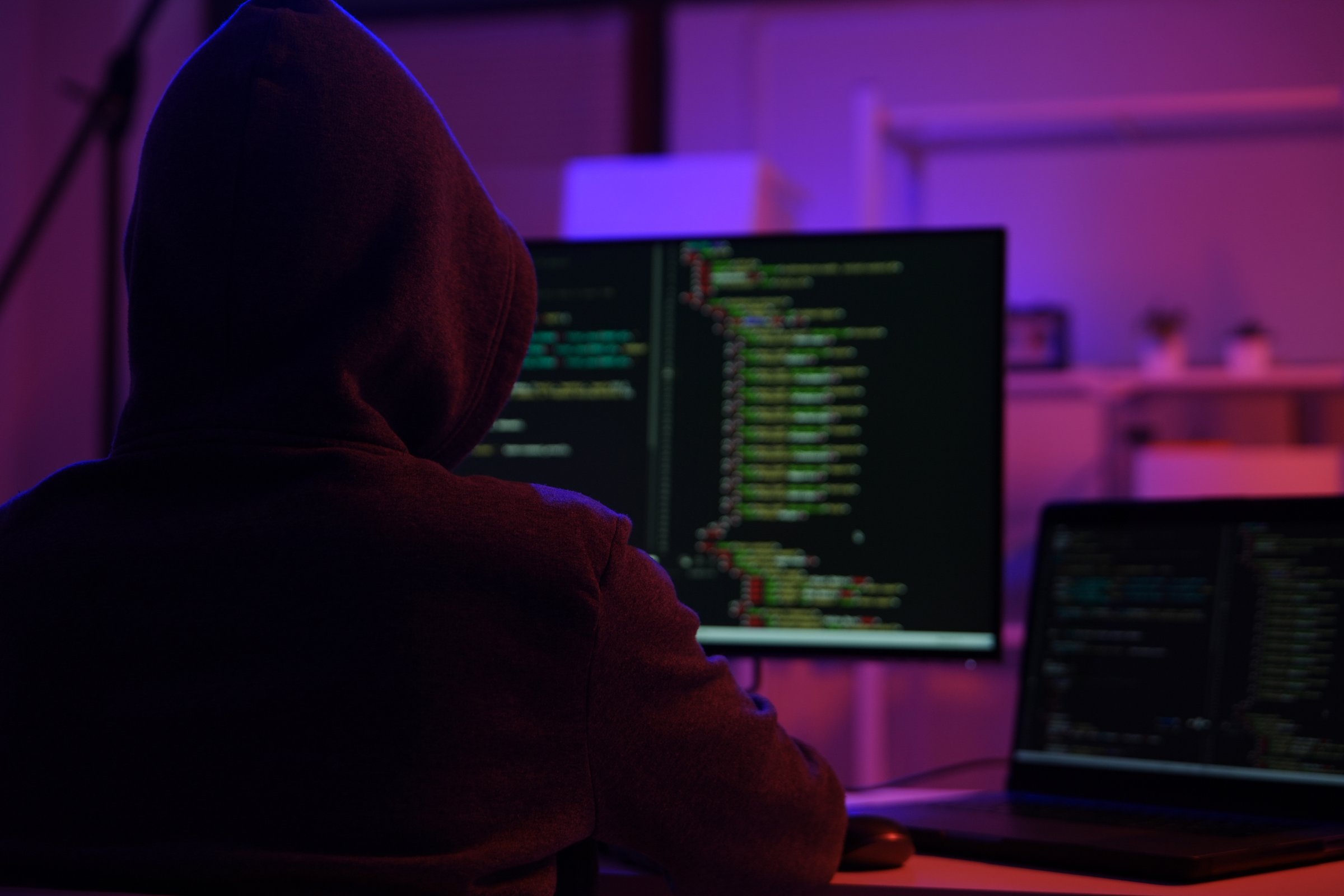 A mysterious hooded hacker working on a computer in a dark room. A concept for cybersecurity, hacking, cybercrime, and data privacy threats.