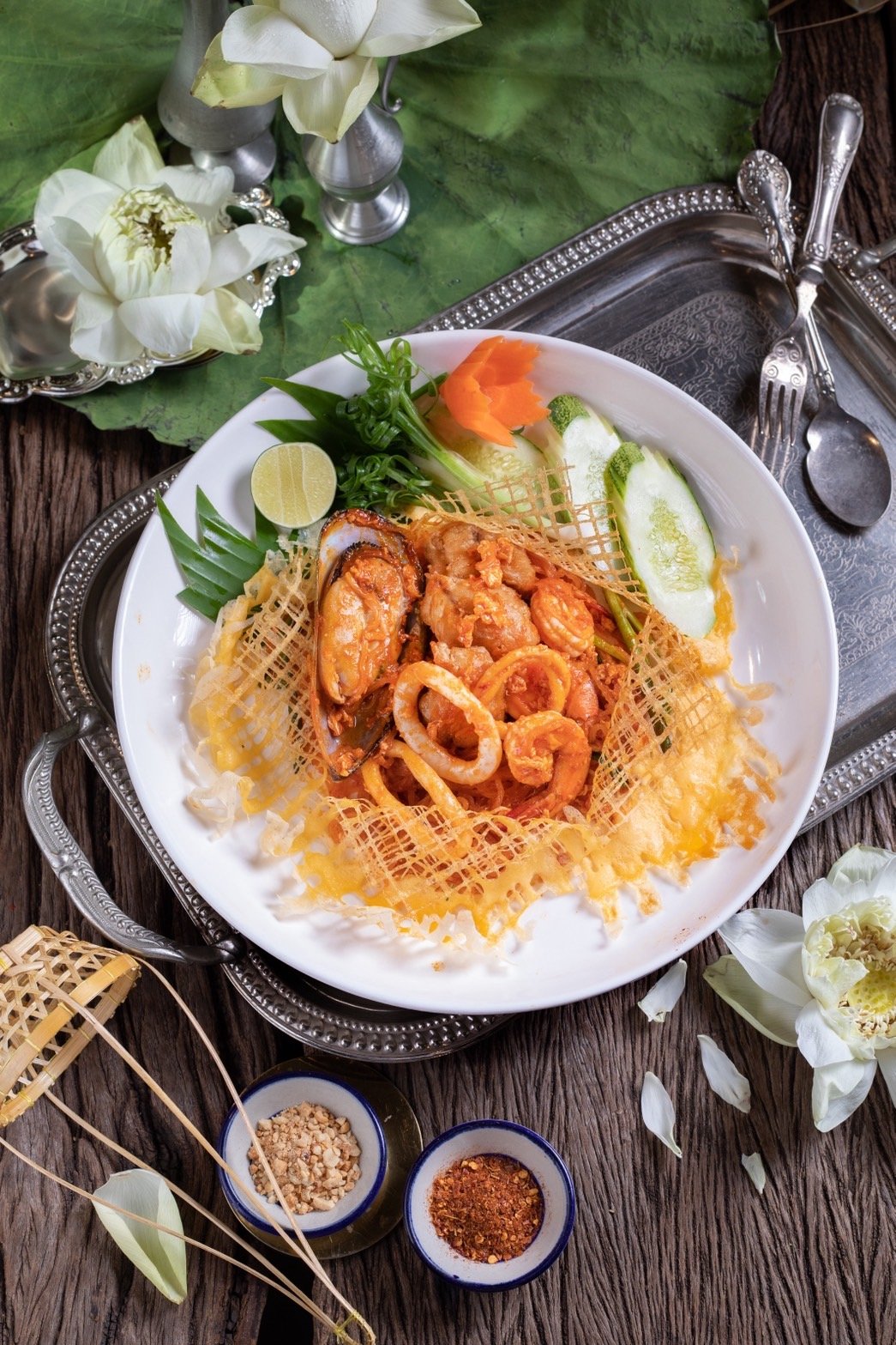 A gourmet dish with seafood, vegetables, and a lattice garnish on a white plate, surrounded by flowers and spices.