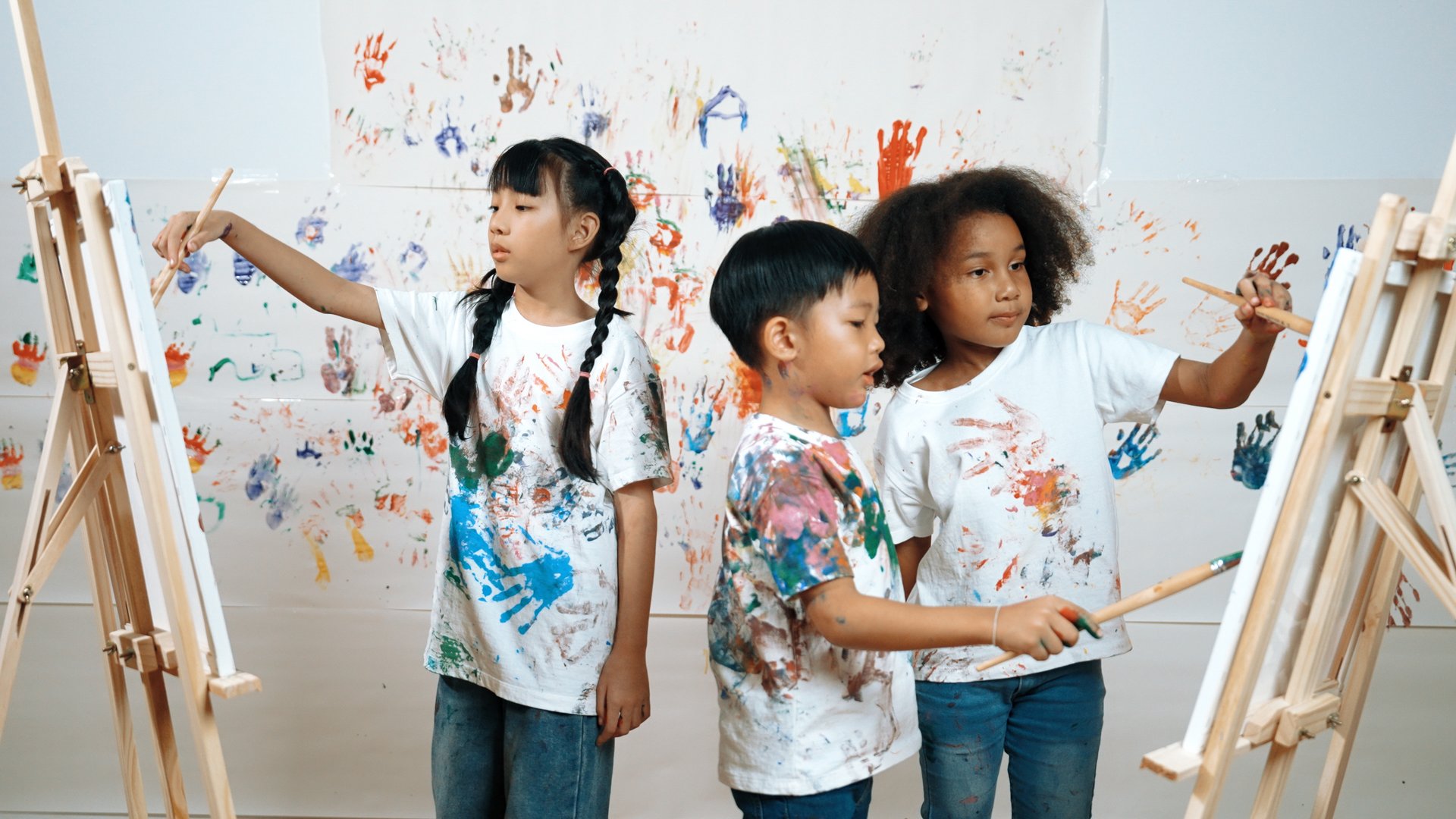 Diverse student painted or draw canvas at stained wall in art lesson. Asian girl wearing white shirt with stained color while standing at stained wall with hand print. Creative activity. Erudition.