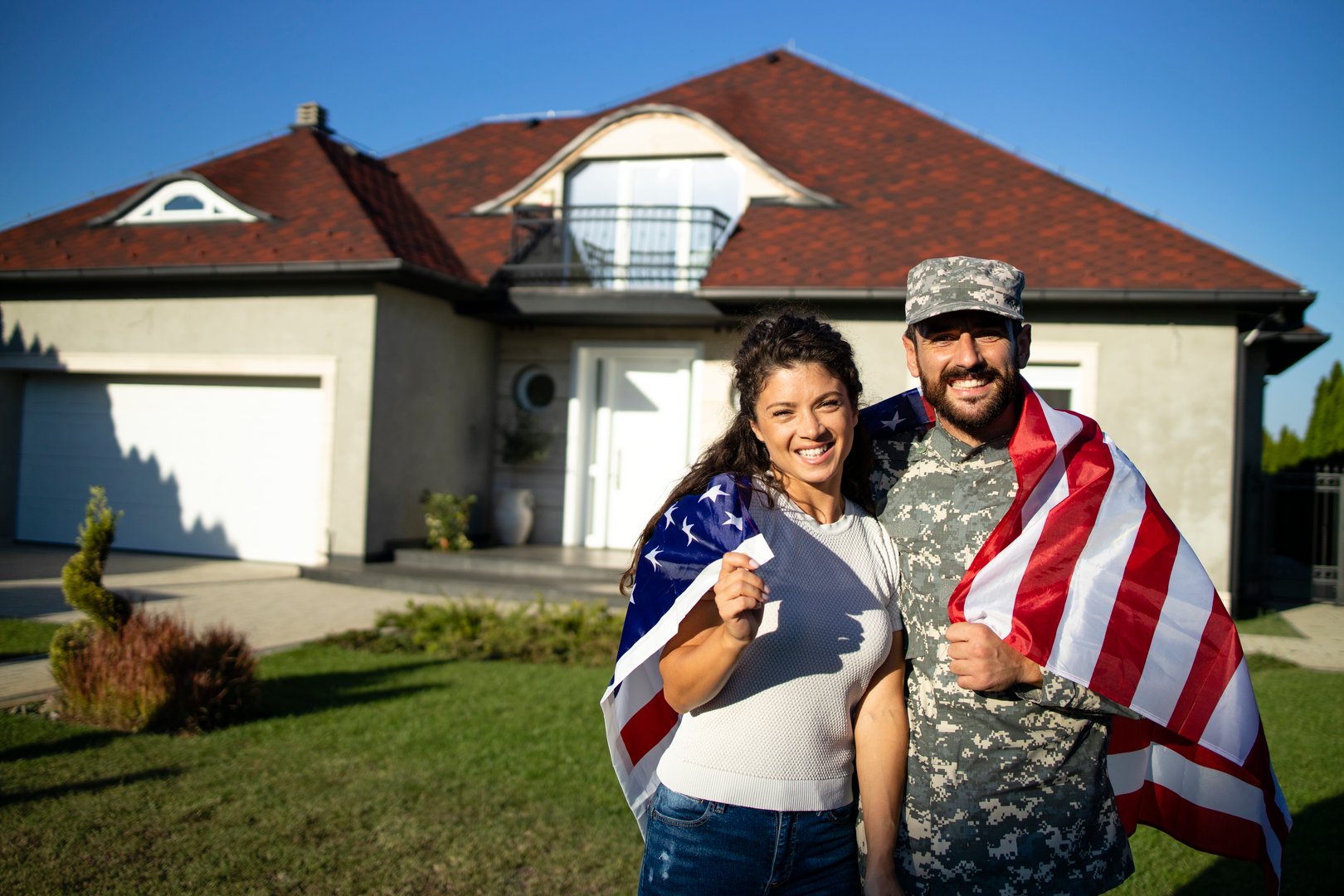 Portrait of husband soldier in uniform on military leave embracing his lovely wife and holding American flag in front of their house.