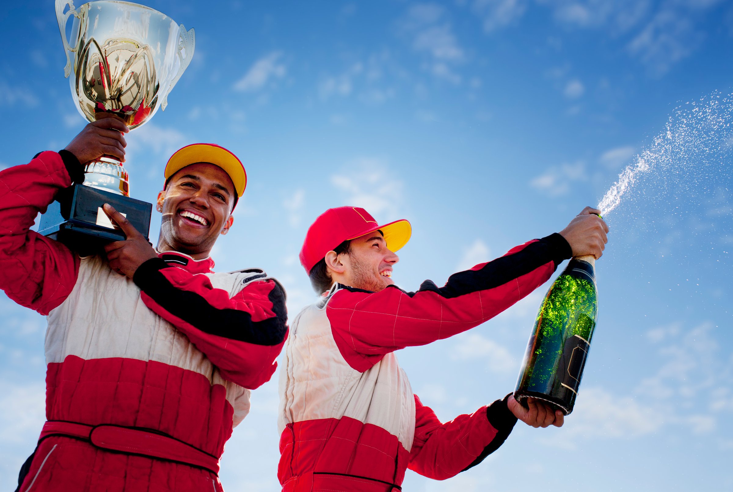 Two racing drivers celebrating victory, wearing red suits and caps, holding a trophy and spraying champagne.