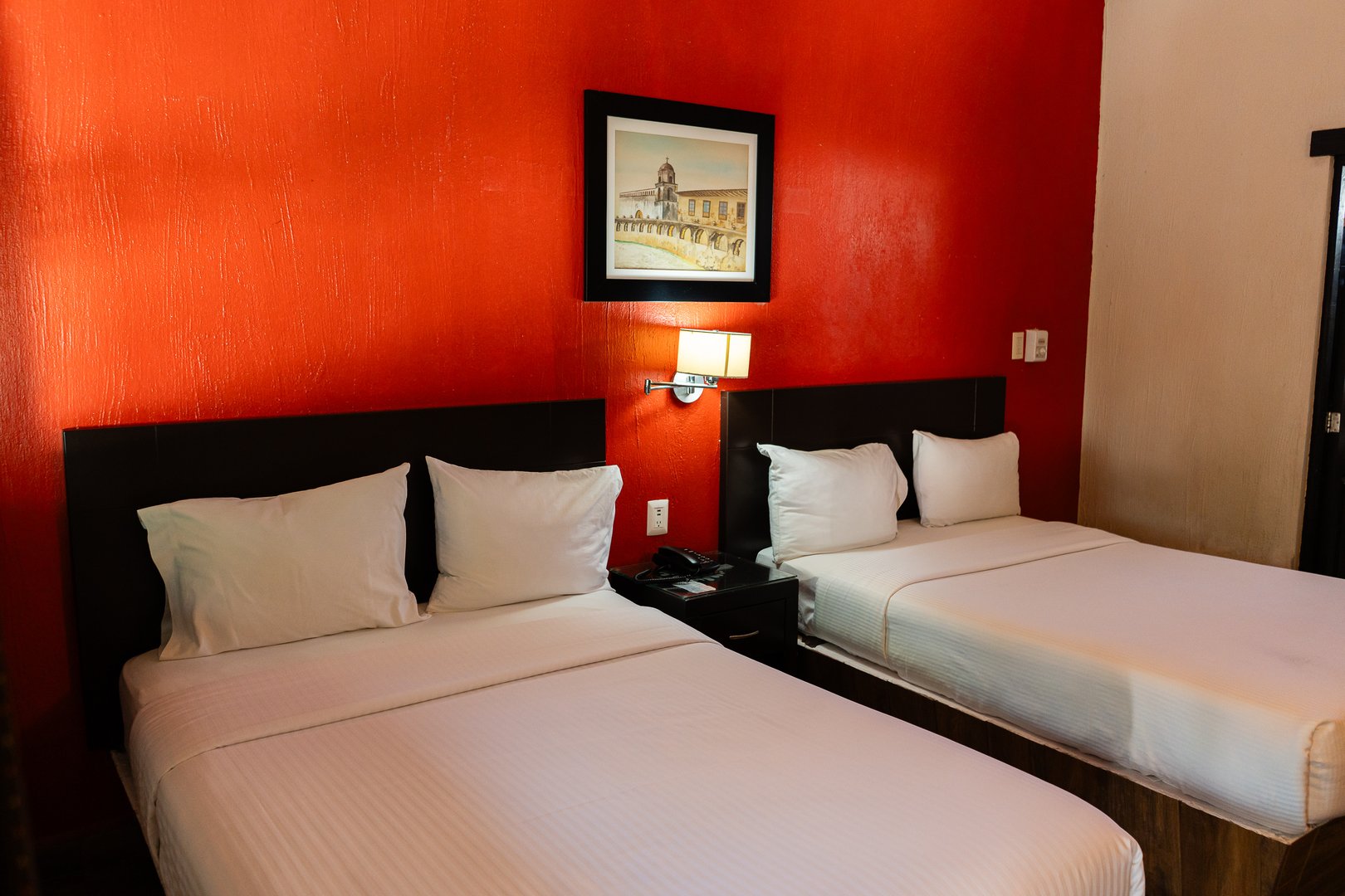 Hotel room with two double beds, white linens, a red accent wall, and framed art above the beds.