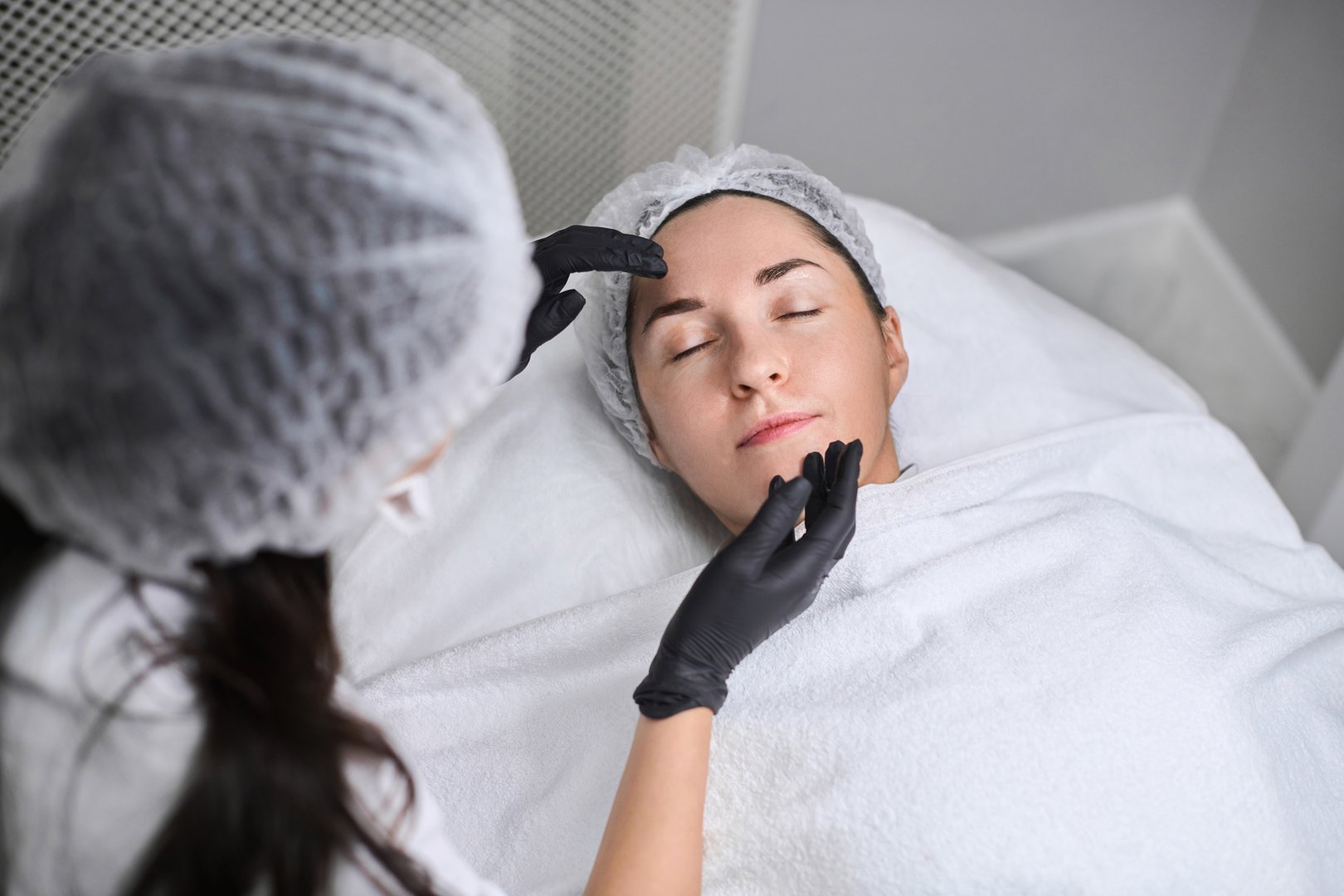 Skincare professional, wearing uniform and gloves, applies facial treatment to relaxed young woman in serene clinic, female beautician wipes client skin of face with cotton pad