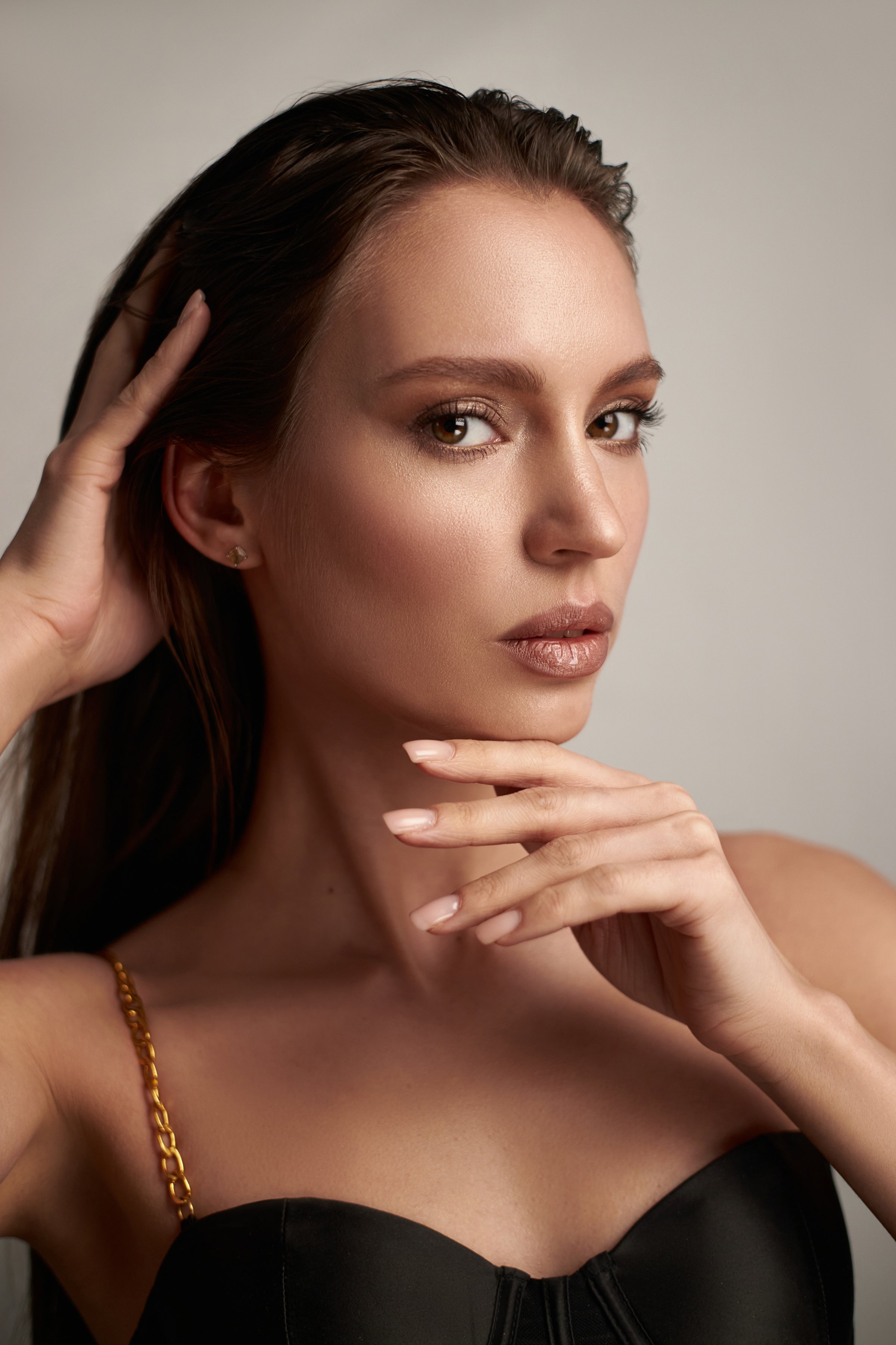 model with long hair strikes a confident pose in a stylish black outfit, using her hands to frame her face while exuding a sense of poise and charm within a well-lit studio.