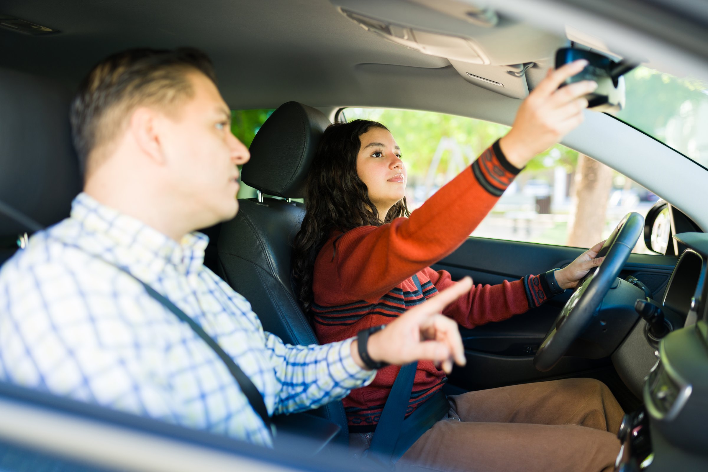 Hispanic teen girl adjusting rearview mirror while learning to drive with her father next to her