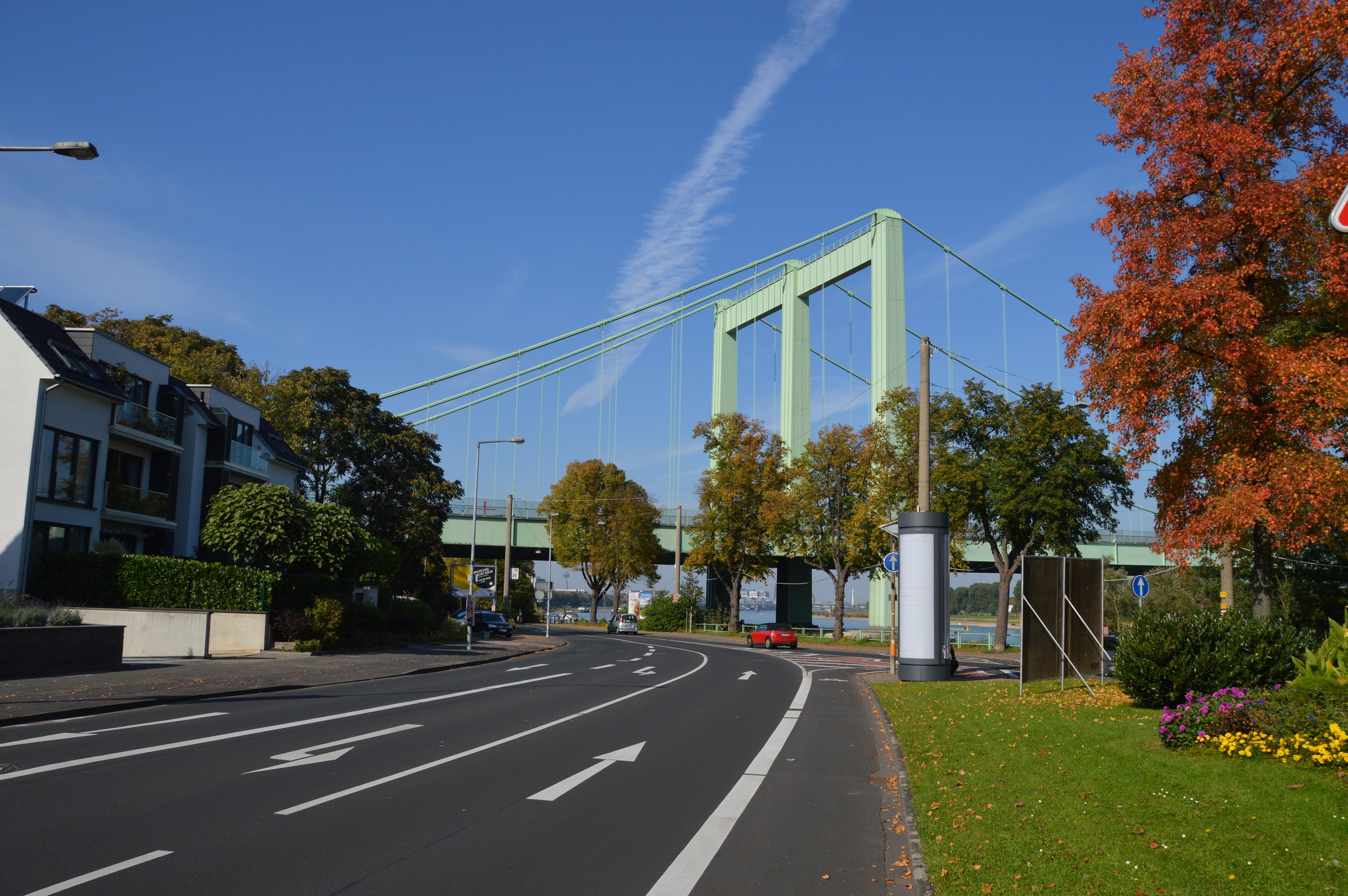 A green suspension bridge over a road with autumn trees on a clear day. Residential buildings are visible on the left.